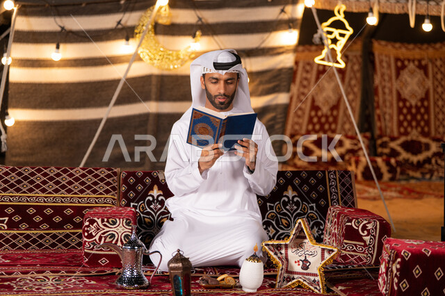 التعبد بتلاوة القران وحفظ وتفسير كلامه، رجل عربي خليجي اماراتي يرتدي الكندوره والغتره البيضاء يحمل المصحف الشريف، الجو الديني الروحاني, العبادة والتقرب إلى رب العالمين، كشته بر شبابيه، التخييم في اجواء رمضانيه، خيمه عربيه مليىْه بديكورات شهر رمضان