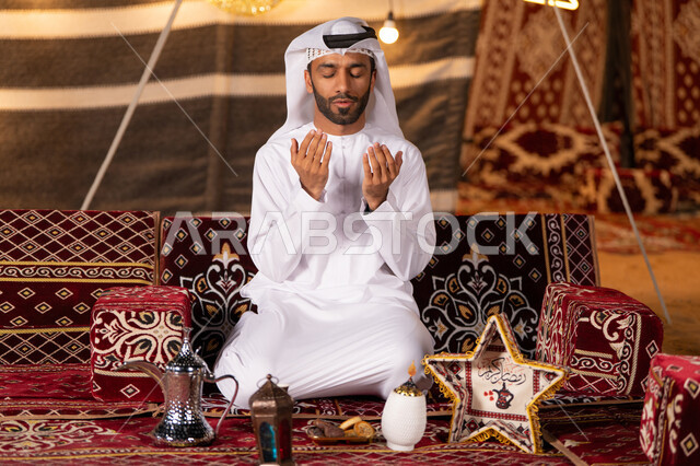 الإكثار من الدعاء في شهر رمضان المبارك، التقرب الى الله من خلال الإجتهاد في العبادة والحفاظ علي الطاعات، شاب عربي خليجي اماراتي يرتدي الكندوره والغتره البيضاء يرفع يديه بالدعاء والتذلل لرب العالمين، خيمه مليىْه بديكورات شهر رمضان الفضيل، التخييم في اجواء وروحانيات وعباده