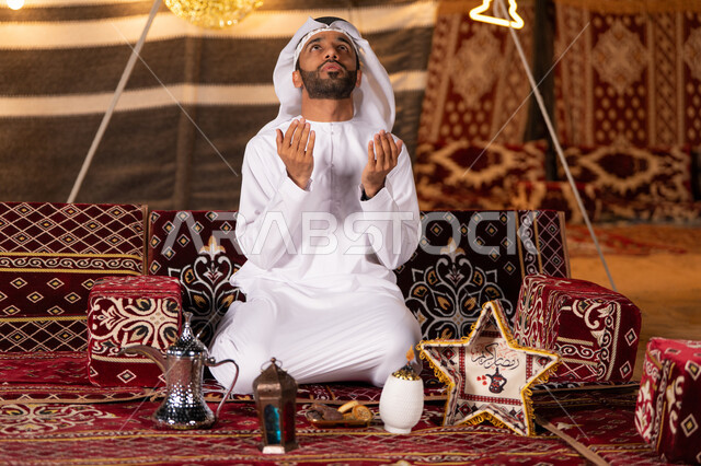 التضرع والخشوع و العبادة, رجل عربي خليجي اماراتي يرتدي الكندوره والغتره البيضاء ينظرالي السماء ويرفع يديه بالدعاء والطلب من الله، ايماءات يد تدل على الدعاء و التقرب من الله، خيمه مليىْه بديكورات شهر رمضان الفضيل، التخييم في اجواء وروحانيات وعباده