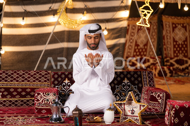الإكثار من الدعاء في شهر رمضان المبارك، التقرب الى الله من خلال الإجتهاد في العبادة والحفاظ علي الطاعات، شاب عربي خليجي اماراتي يرتدي الكندوره والغتره البيضاء يرفع يديه بالدعاء والتذلل لرب العالمين، خيمه مليىْه بديكورات شهر رمضان الفضيل، التخييم في اجواء وروحانيات وعباده