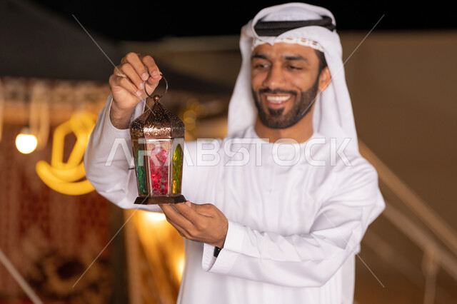 فانوس معدني مزخرف ملون, الاستعداد لاستقبال الشهر الفضيل، شاب عربي خليجي اماراتي يرتدي الكندوره والغتره البيضاء مبتسم يحمل فانوس معدني تقليدي, الأجواء الروحانية في الأيام الفضيلة, زينة و ديكورات و أنوار الاحتفالات بالمناسبات و الأعياد الدينية الإسلامية, البهجة بقدوم رمضان المبارك