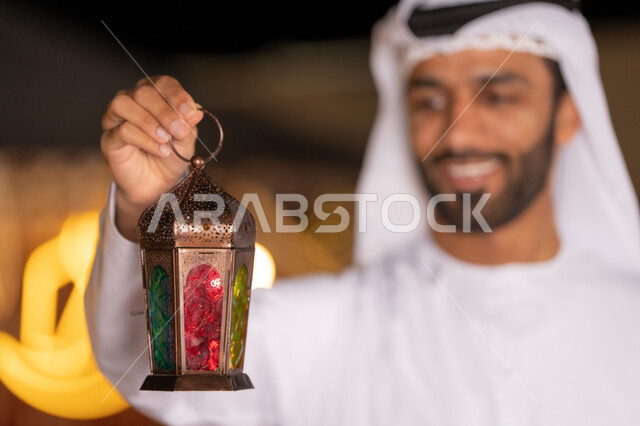 فانوس معدني مزخرف ملون, الاستعداد لاستقبال الشهر الفضيل، شاب عربي خليجي اماراتي يرتدي الكندوره والغتره البيضاء مبتسم يحمل فانوس معدني تقليدي, الأجواء الروحانية في الأيام الفضيلة, زينة و ديكورات و أنوار الاحتفالات بالمناسبات و الأعياد الدينية الإسلامية, البهجة بقدوم رمضان المبارك
