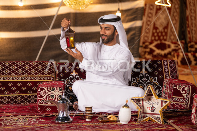 ديكورات الأجواء الرمضانية الروحانية، مجموعة متنوعة من الفوانيس للاحتفال بقدوم شهر رمضان، شاب عربي خليجي اماراتي يرتدي الكندوره والغتره البيضاء مبتسم يمسك فانوس رمضان ، كشته وجلسه بر، التخييم الليلي الهادىْ في صحاري الامارات، تزيين خيمه شعبيه تراثيه