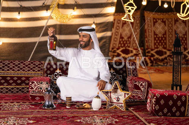 Spiritual Ramadan atmosphere decorations, a variety of lanterns to celebrate the arrival of Ramadan, a young Emirati Gulf Arab man wearing a white kandura and ghutra, smiling, holding a Ramadan lantern, a kashta and a desert session, a quiet night camping in the UAE desert, decorating a traditional traditional tent