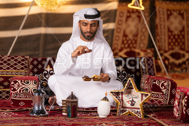 كسر الصيام بتناول التمر والحلوي الرمضانيه اثناء التخييم، مجموعة من الفوانيس للاحتفال بقدوم شهر رمضان، شاب عربي خليجي اماراتي يرتدي الكندوره والغتره البيضاء مبتسم يمسك حبه من التمر، كشته بر، خيمه مضيىْه في الليل الهادىْ، عادات وتقاليد امراتيه في اكرام الضيف