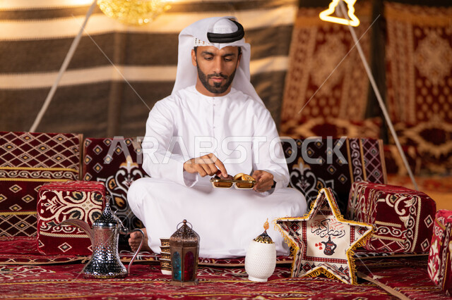 كسر الصيام بتناول التمر والحلوي الرمضانيه اثناء التخييم، مجموعة من الفوانيس للاحتفال بقدوم شهر رمضان، شاب عربي خليجي اماراتي يرتدي الكندوره والغتره البيضاء مبتسم يمسك حبه من التمر، كشته بر، خيمه مضيىْه في الليل الهادىْ، عادات وتقاليد امراتيه في اكرام الضيف