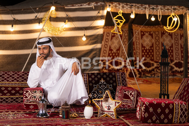 تناول القهوه العربيه الفاخره اثناء التخييم، تزيين الخيمه بالفوانيس للاحتفال بقدوم شهر رمضان، شاب عربي خليجي اماراتي يرتدي الكندوره والغتره البيضاء يمسك كوب من المشروبات الساخنه، كشته بر، خيمه مضيىْه في الليل الهادىْ، عادات وتقاليد امراتيه في اكرام الضيف