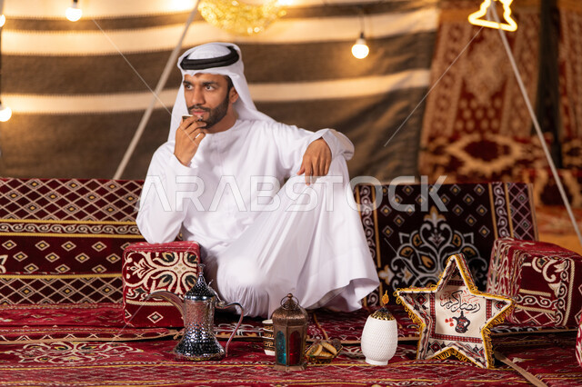 تناول القهوه العربيه الفاخره اثناء التخييم، تزيين الخيمه بالفوانيس للاحتفال بقدوم شهر رمضان، شاب عربي خليجي اماراتي يرتدي الكندوره والغتره البيضاء يمسك كوب من المشروبات الساخنه، كشته بر، خيمه مضيىْه في الليل الهادىْ، عادات وتقاليد امراتيه في اكرام الضيف