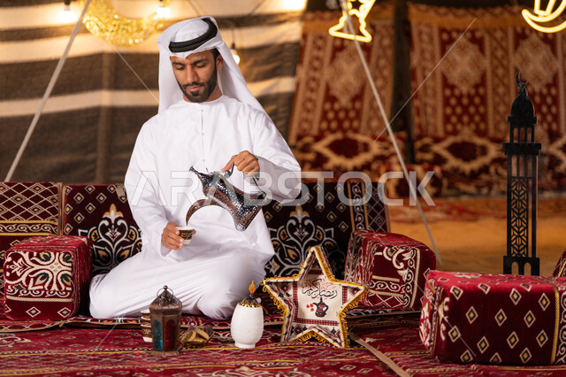 الكرم وحسن الضيافة والإستقبال عند العرب، ديكورات الأجواء الرمضانية الروحانية، مجموعة متنوعة من الفوانيس للاحتفال بقدوم شهر رمضان، شاب عربي خليجي اماراتي يرتدي الكندوره والغتره البيضاء يسكب القهوه الساخنه، كشته وجلسه بر، التخييم وسط صحراء الامارات