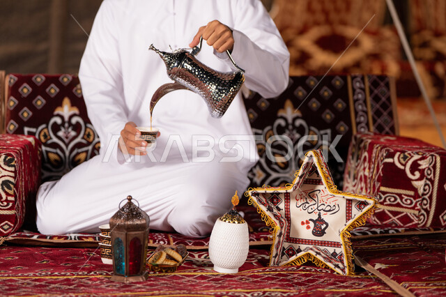 تقديم القهوه بالطريقة التقليدية الاماراتية, مفهوم الكرم وحسن الاستقبال, صورة مقربة لشاب عربي خليجي اماراتي يرتدي الغتره البيضاء يسكب القهوه من الدله النحاسيه, ديكورات وزينه شهر رمضان الكريم، التخييم الهاديْ ليلا، كشته بريه شبابيه، خيمه عربيه تراثيه