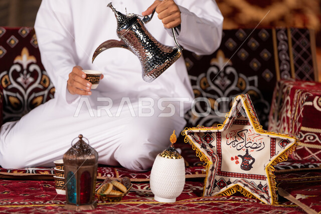 تقديم القهوه بالطريقة التقليدية الاماراتية, مفهوم الكرم وحسن الاستقبال, صورة مقربة لشاب عربي خليجي اماراتي يرتدي الغتره البيضاء يسكب القهوه من الدله النحاسيه, ديكورات وزينه شهر رمضان الكريم، التخييم الهاديْ ليلا، كشته بريه شبابيه، خيمه عربيه تراثيه