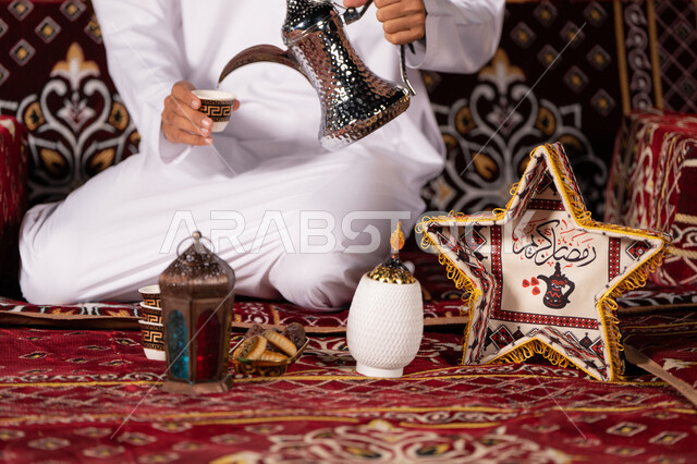 تقديم القهوه بالطريقة التقليدية الاماراتية, مفهوم الكرم وحسن الاستقبال, صورة مقربة لشاب عربي خليجي اماراتي يرتدي الغتره البيضاء يسكب القهوه من الدله النحاسيه, ديكورات وزينه شهر رمضان الكريم، التخييم الهاديْ ليلا، كشته بريه شبابيه، خيمه عربيه تراثيه