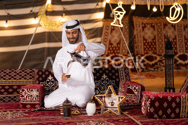 عادات وتقاليد أصيلة في الاستقبال وتقديم المشروبات الساخنه، شاب عربي خليجي اماراتي مبتسم يرتدي الكندوره والغتره البيضاء يقوم بصب القهوة من الدلة النحاسية في فنجان داخل خيمة شعبية تراثية مضيىْه بديكورات شهر رمضان الكريم، كشه بر مساىْيه، التخييم ليلا