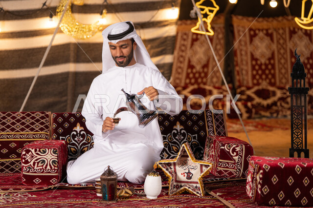 عادات وتقاليد أصيلة في الاستقبال وتقديم المشروبات الساخنه، شاب عربي خليجي اماراتي مبتسم يرتدي الكندوره والغتره البيضاء يقوم بصب القهوة من الدلة النحاسية في فنجان داخل خيمة شعبية تراثية مضيىْه بديكورات شهر رمضان الكريم، كشه بر مساىْيه، التخييم ليلا