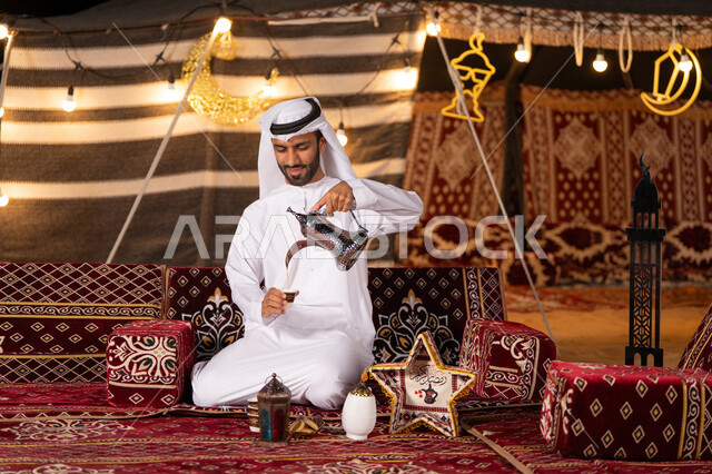 عادات وتقاليد أصيلة في الاستقبال وتقديم المشروبات الساخنه، شاب عربي خليجي اماراتي مبتسم يرتدي الكندوره والغتره البيضاء يقوم بصب القهوة من الدلة النحاسية في فنجان داخل خيمة شعبية تراثية مضيىْه بديكورات شهر رمضان الكريم، كشه بر مساىْيه، التخييم ليلا