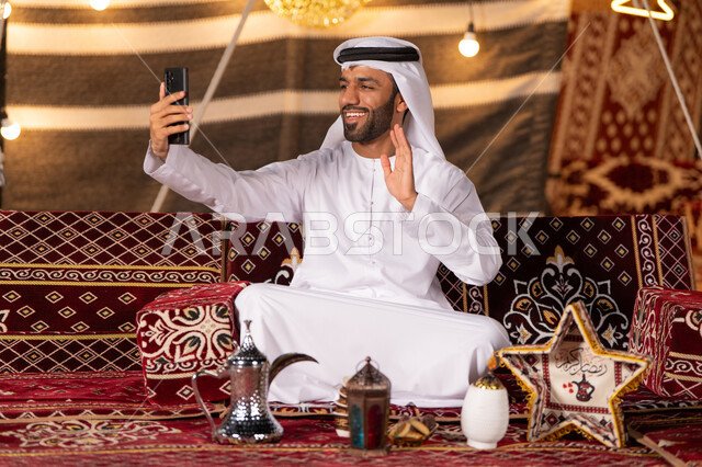 بث فيديومباشر، شاب عربي خليجي اماراتي مبتسم يرتدي الكندوره والغتره البيضاء يستخدم الهاتف المحمول، التخييم في اجواء شهر رمضان المبارك، كشته بريه شبابيه في صحاري الامارات، خيمه عربيه شعبيه مضيىْه بديكورات رمضانيه، ايماءات الابتسامه والترحيب