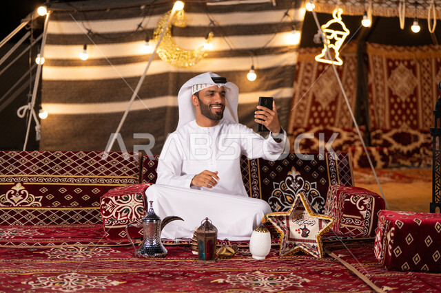 تزيين خيمة عربيه بديكورات وزينه رمضانيه في صحاري الامارات، رجل عربي خليجي اماراتي مبتسم يرتدي الكندوره والغتره البيضاء يستخدم الهاتف المحمول، التخييم الليلي في الصحراء، استخدام التقنيات والوسائل الحديثة، كشته بريه شبابيه،الاستمتاع بشهررمضان الكريم