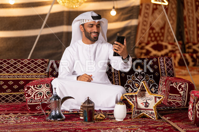 استخدام وسائل التواصل الاجتماعي، بث فيديو مباشر، رحلة تخييم في الصحراءليلا، خيمه بريه عربية شعبية، كشتة بر مسائية، اجواء شهررمضان المبارك، شاب عربي خليجي اماراتي مبتسم يرتدي الكندوره والغتره البيضاء يستخدم الهاتف المحمول، ديكورات وزينه رمضانيه