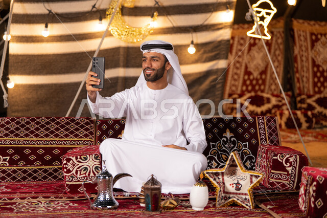 التقاط صوره سيلفي تذكاريه, شاب عربيى خليجي اماراتي مبتسم يرتدي الكندوره والغتره البيضاء يجلس في خيمه عربيه تقليديه, التخييم الليلي في صحراء الامارات, خيمه مضيىْه بديكورات رمضانيه, كشتة بر مسائية, ديكورات وزينه شهر رمضان المبارك
