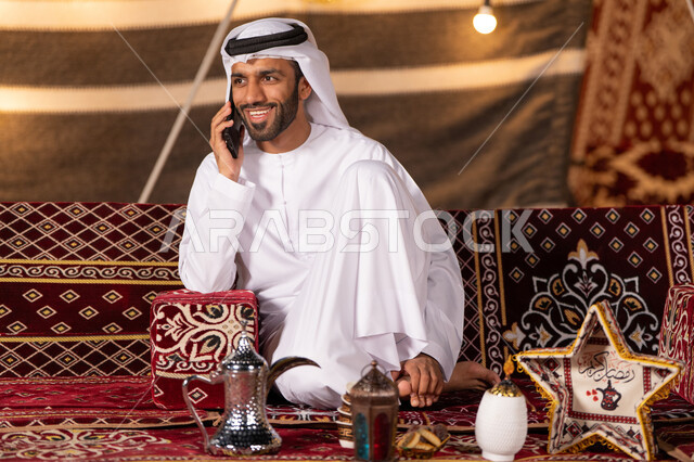 قضاء وقت ممتع في التخييم الليلي الهادئ, رجل عربي خليجي اماراتي مبتسم يرتدي الكندوره والغتره البيضاء يجري مكاملة هاتفية ، كشتة بر شبابية, خيمه مضيىْه بديكورات وزينه رمضانيه, الاستمتاع بشهر رمضان الكريم