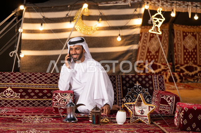 قضاء وقت ممتع في التخييم الليلي الهادئ, رجل عربي خليجي اماراتي مبتسم يرتدي الكندوره والغتره البيضاء يجري مكاملة هاتفية ، كشتة بر شبابية, خيمه مضيىْه بديكورات وزينه رمضانيه, الاستمتاع بشهر رمضان الكريم