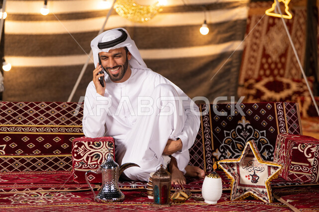 التخييم في صحاري الامارات ليلا, شاب عربي خليجي اماراتي مبتسم يرتدي الكندوره والغتره البيضاء يجري مكالمة هاتفية, ايماءات الفرح والسرور, كشته بر شبابيه, خيمه شعبيه تقليديه مضيئه بزينة شهر رمضان