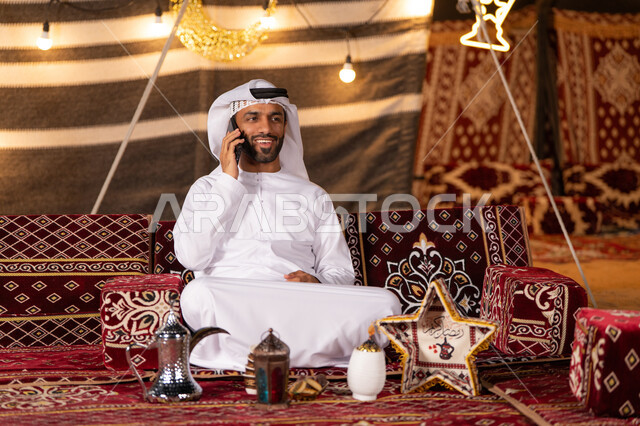 التخييم في شهر رمضان المبارك, شاب عربي خليجي اماراتي مبتسم  يجري مكالمة هاتفية, كشتة و جلسة بر شبابية, خيمه مضيىْه بزينه وديكورات رمضانيه