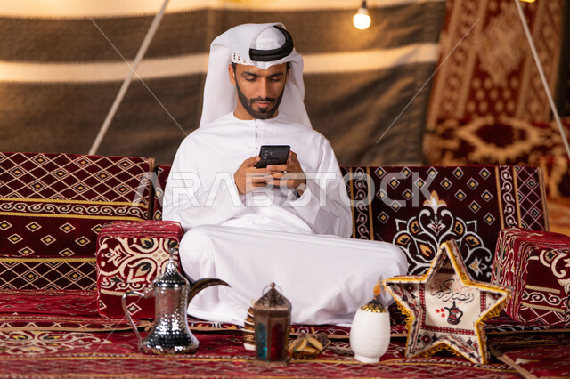 An Emirati Gulf Arab man wearing a white kandura and ghutra sitting in a traditional tent looking at his phone, a youth evening outing, Ramadan decorations, camping at night in the UAE desertكش