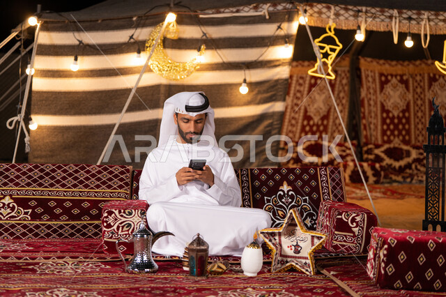 زينه وديكورات شهر رمضان المبارك في خيمه تقليديه في صحراء الامارات, كشتة بر شبابية, رجل عربي خليجي اماراتي يرتدي الكندوره والغتره البيضاء يجلس في خيمه تقليديه ينظر الي الهاتف, اجواء التخييم في شهر رمضان المبارك