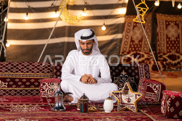 الأصالة والعراقة والتراث الاماراتي, استعداد المسلمين لاستقبال شهر رمضان, شاب اماراتي يرتدي الكندوره والغتره البيضاء يجلس بأيدي متماسكه وينظر الي الاسفل, كشته بر شبابية, التخييم الليلي الهاديْ فى صحراء الامارات, خيمه مضيىْه بديكورات رمضانيه