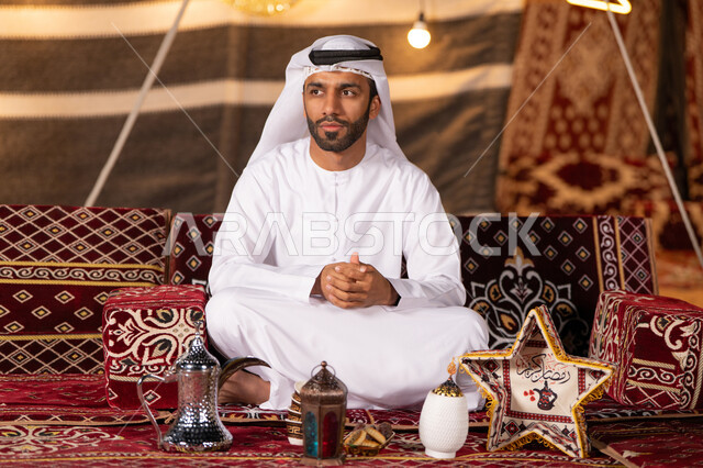 A young Emirati Gulf Arab man wearing a white kandura and ghutra sitting with his hands clasped, a traditional Bedouin tent illuminated with Ramadan decorations, a quiet night camping in the UAE desert, a youth camp in the desert, gestures of absent-mindedness