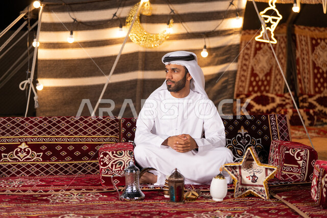 خيمه شعبيه مضيىْه بديكورات رمضانيه، رجل عربي خليجي اماراتي يرتدي الكندوره والغترة البيضاء يجلس بأيدي متشبكه، كشتة بر في صحاري الامارات,  التخييم في الليل الهاديْ