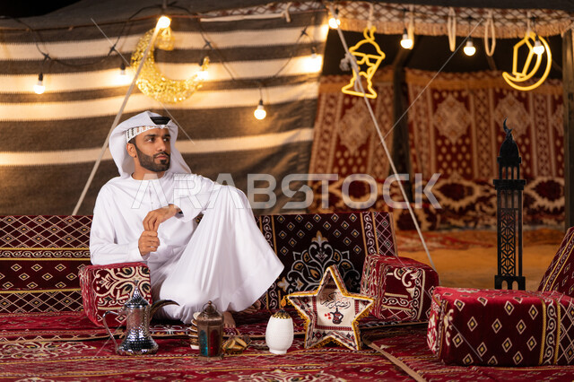 تزيين الخيمه بفوانيس وديكورات رمضان, ديكورات وزينه شهر رمضان المبارك, شاب عربي خليجي اماراتي يرتدي الكندوره والغتره البيضاء يجلس بأيدي متماسكه في خيمه عربيه شعبيه في صحراء الامارات,  كشتة بر شبابية, التخييم في الليل الهادىْ