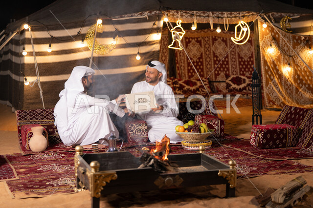 ديكورات وزينه رمضانيه, صديقان عربيان اماراتيان خليجيان يرتديان الكندوره والغتره يجلسان في خيمه عربية تقليدية امام شعلة الحطب, أجواء ليلية هادىْه, كشتة بر شبابية, تقديم الهدية في المناسبات الدينية, كرم الضيافة وحسن الاستقبال, التخييم في شهر رمضان المبارك