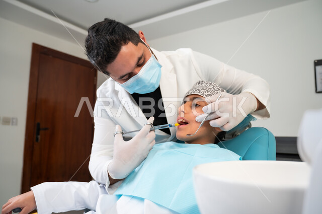 استخدام ابرة التخدير لتخفيف الألم، ولد عربي خليجي عماني يرتدي الزي التقليدي يجلس في العيادة السنية يتلقى العلاج اللازم، طبيب أسنان يرتدي المعطف والقفازات والكمامة الطبية يقوم بحقن البنج الموضعي، زيارة العيادات الصحية واتخاذ الإجراءات اللازمة