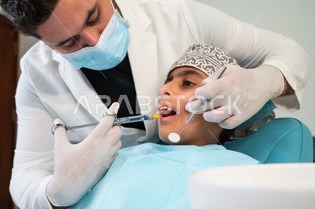 زيارة العيادات الصحية بشكل دوري، استخدام ابرة التخدير لتخفيف الألم، صورة مقربة لولد عربي خليجي عماني يرتدي الزي التقليدي يجلس في العيادة السنية يتلقى العلاج اللازم، طبيب أسنان يرتدي المعطف والقفازات والكمامة الطبية يقوم بحقن البنج الموضعي