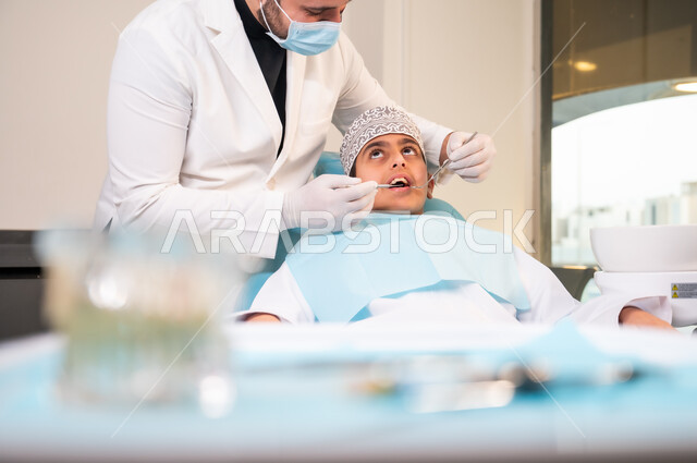 الاستعداد لتلقي العلاج اللازم، المحافظة على الصحة الفموية، التأكد من سلامة الأسنان واللثة، المعدات والأدوات المعقمة، طبيب عربي خليجي عماني يرتدي المعطف والقفازات والكمامة الطبية يقوم بفحص المريض، صورة مقربة لولد يرتدي الزي التقليدي يجلس في العيادة السنية