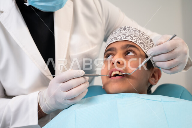 التأكد من سلامة أسنان ولثة المريض، استعمال المعدات والأدوات النظيفة والمعقمة، صورة مقربة لولد عربي خليجي عماني يرتدي الزي التقليدي ينظر إلى طبيب يرتدي القفازات الطبية يمسك بيده أداة الفحص بايماءات الاطمئنان والسرور، تقديم العلاج اللازم في المراكز الصحية