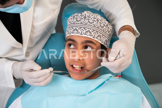 Preparing to receive the necessary treatment, maintaining oral health, ensuring the safety of teeth and gums, sterile equipment and tools, an Arab Gulf Omani doctor wearing a coat, gloves and a medical mask examining a patient, a close-up of a boy wearing traditional clothing sitting in the dental clinic