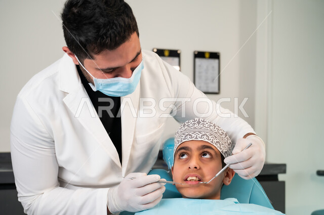 التأكد من سلامة أسنان ولثة المريض، استعمال المعدات والأدوات النظيفة والمعقمة، صورة مقربة لولد عربي خليجي عماني يرتدي الزي التقليدي ينظر إلى طبيب يرتدي القفازات الطبية يمسك بيده أداة الفحص بايماءات الاطمئنان والسرور، تقديم العلاج اللازم في المراكز الصحية