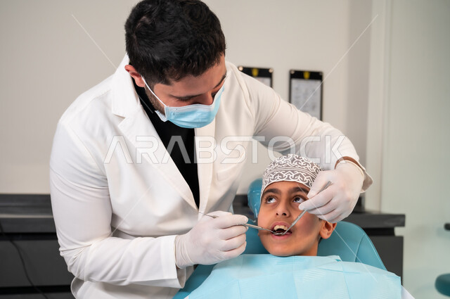 طب وجراحة الفم والفكين، الزيارة الدورية للمراكز الصحية، عيادة أسنان خاصة، استخدام المعدات والأدوات النظيفة والمعقمة، طبيب يرتدي المعطف والقفازات الطبية يفحص فم المريض، صورة مقربة لولد عربي خليجي عماني يرتدي الزي التقليدي يجلس على كرسي العلاج