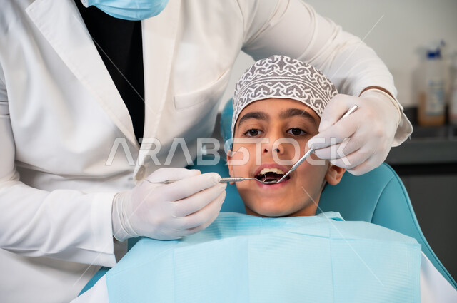 عيادة أسنان خاصة، استخدام المعدات والأدوات النظيفة والمعقمة، الزيارة الدورية للمراكز الصحية، طبيب يرتدي المعطف والقفازات الطبية يفحص فم المريض، صورة مقربة لولد عربي خليجي عماني يرتدي الزي التقليدي يجلس على كرسي العلاج