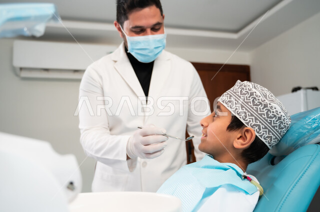 تقديم العلاج اللازم في المراكز الصحية، طب وجراحة الفم والفكين، استخدام المعدات والأدوات النظيفة والمعقمة، طبيب يرتدي المعطف والقفازات الطبية يقوم بفحص فم الفتى المريض، صورة مقربة لولد عربي خليجي عماني يرتدي الزي التقليدي يجلس في العيادة السنية