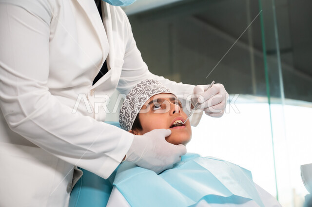طب وجراحة الفم والفكين، الزيارة الدورية للمراكز الصحية، عيادة أسنان خاصة، استخدام المعدات والأدوات النظيفة والمعقمة، طبيب يرتدي المعطف والقفازات الطبية يفحص فم المريض، صورة مقربة لولد عربي خليجي عماني يرتدي الزي التقليدي يجلس على كرسي العلاج