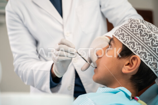 التأكد من سلامة الأسنان واللثة باستخدام المعدات والأدوات المعقمة، الزيارة الدورية للعيادات الصحية، طبيب يرتدي المعطف والقفازات الطبية يقوم بفحص فم المريض، صورة مقربة من الجانب لولد عربي خليجي عماني يرتدي الكمه يجلس في العيادة السنية يتلقى العلاج اللازم
