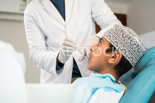 المعدات والأدوات المعقمة، الزيارة الدورية للعيادات الصحية، المحافظة على الصحة الفموية، طبيب أسنان يرتدي المعطف والقفازات الطبية يقوم بفحص فم المريض، صورة مقربة من الجانب لولد عربي خليجي عماني يرتدي الكمه يجلس في العيادة السنية يتلقى العلاج اللازم