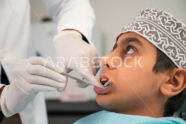 Sterilized equipment and tools, regular visit to health clinics, maintaining oral health, dentist wearing medical coat and gloves examining patient's mouth, close-up side view of an Arab Gulf Omani boy wearing a mask sitting in the dental clinic receiving necessary treatment