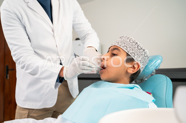 الزيارة الدورية للعيادات الصحية لاتخاذ الإجراءات اللازمة، المحافظة على الصحة الفموية، الأدوات والمعدات المعقمة، طبيب أسنان يرتدي المعطف والقفازات الطبية يقوم بفحص فم المريض، صورة مقربة لولد عربي خليجي عماني يرتدي الكمه يجلس في العيادة السنية يتلقى العلاج اللازم