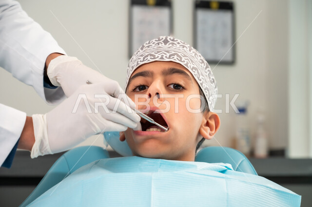 النظر إلى الكاميرا أثناء الفحص الطبي، التأكد من سلامة الأسنان واللثة باستخدام المعدات والأدوات المعقمة، الزيارة الدورية للعيادة الصحية، طبيب يرتدي المعطف والقفازات الطبية يفحص فم المريض، صورة مقربة لولد عربي خليجي عماني يرتدي الزي التقليدي يجلس في العيادة السنية
