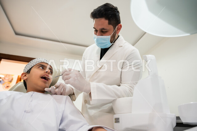 المحافظة على الصحة الفموية، الأدوات والمعدات المعقمة، زيارة العيادات الصحية لاتخاذ الإجراءات اللازمة، طبيب أسنان يرتدي المعطف والقفازات والكمامة الطبية يمسك بيده أداة الفحص، صورة مقربة لولد عربي خليجي عماني يرتدي الكمه يجلس في العيادة السنية يتلقى العلاج اللازم